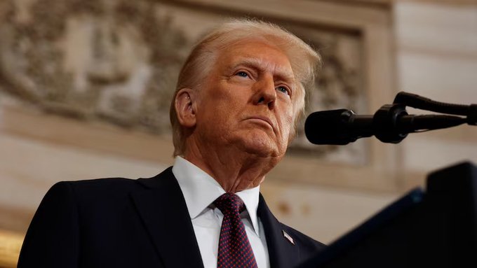 A close-up portrait of Donald Trump in a dark suit and red tie standing at a podium with microphones, looking up and to the side with a serious demeanor.