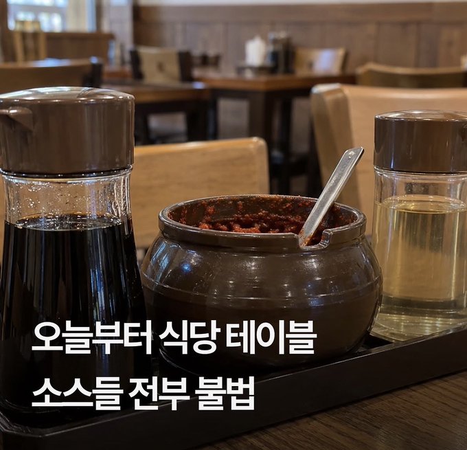 A condiment set on a restaurant table featuring a dark ceramic pot with red sauce and two glass dispensers, with Korean text reading '오늘부터 식당 테이블 소스들 전부 불법'.