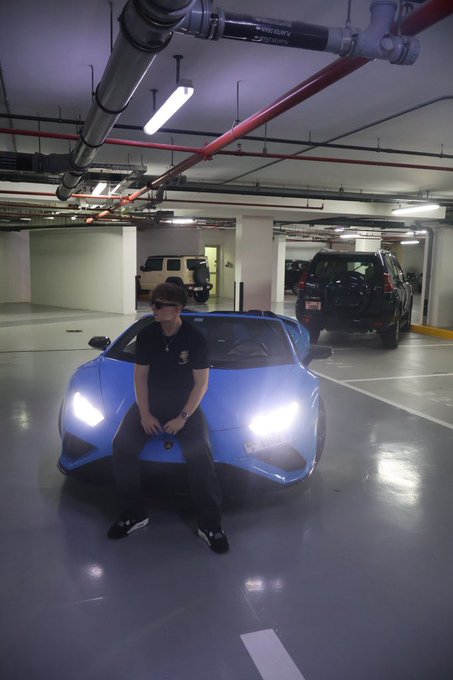 A young man in sunglasses and a dark t-shirt poses sitting on the hood of a sleek blue McLaren sports car with its headlights on, parked in an underground garage. The caption "GN 💙" suggests a "good night" post, likely sharing an evening showcase of his luxurious ride.