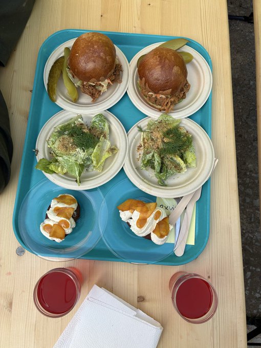 An overhead shot of a blue tray holding a meal: two pulled pork sliders with pickles, two Caesar salads, two small dishes of dessert with meringue, and two small cups of red juice.