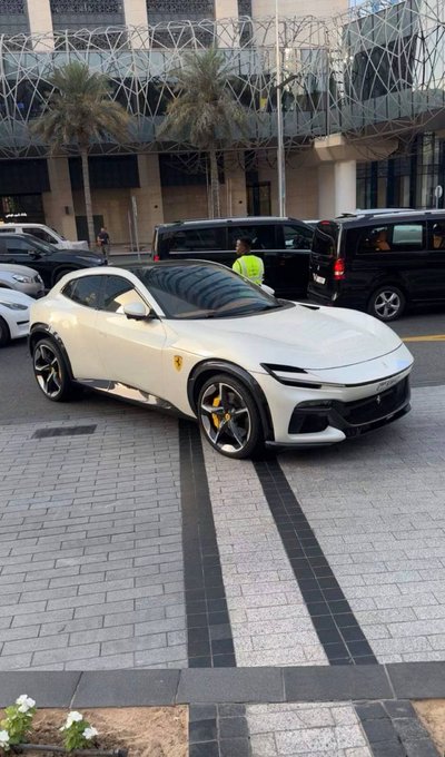 A white Ferrari Purosangue SUV is parked on a paved street in front of a modern building with palm trees, featuring distinctive black wheels with yellow brake calipers.
