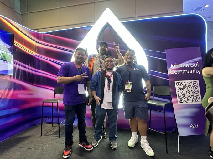 Three men pose for a photo at a Sui blockchain event, standing in front of a glowing Sui logo with a banner reading 'Join the Sui Community' and a QR code.