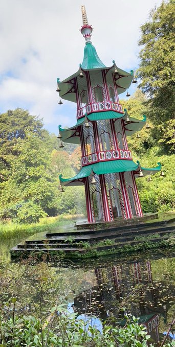 A multi-tiered, ornate Chinese-style pagoda with a green roof and red framing stands on stone near a body of water that reflects its image, surrounded by lush green trees.