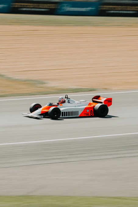 An orange and white Formula One-style race car with the number 3 speeds along a track, with a motion-blurred sandy background conveying high speed.