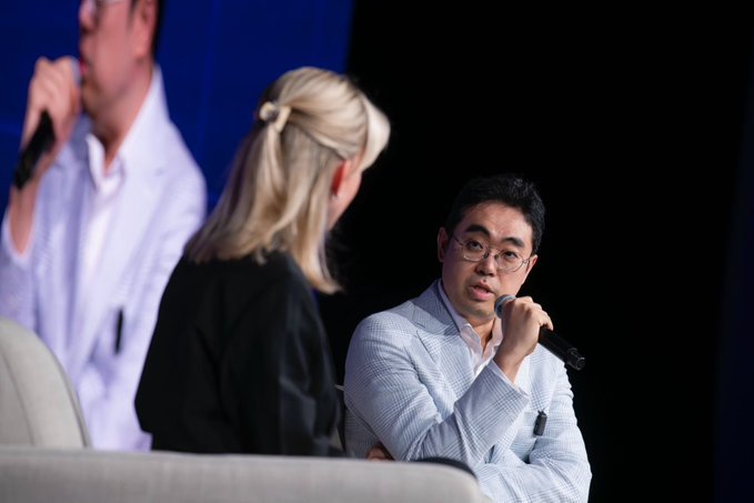 A video conference call shows a man wearing glasses holding a microphone, while another person appears to be engaged in conversation with him or observing something in front of them. There are two people present in the scene.