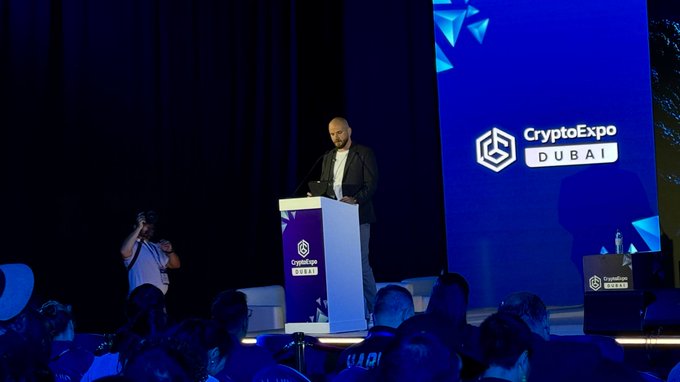 A bald man with a beard stands at a podium on stage at the 'CryptoExpo Dubai' conference, addressing an audience with blue lighting and event branding visible.