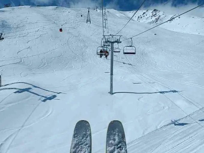 A point-of-view shot from a chairlift showing skis pointing down a snowy mountain slope with other chairlifts visible ahead.