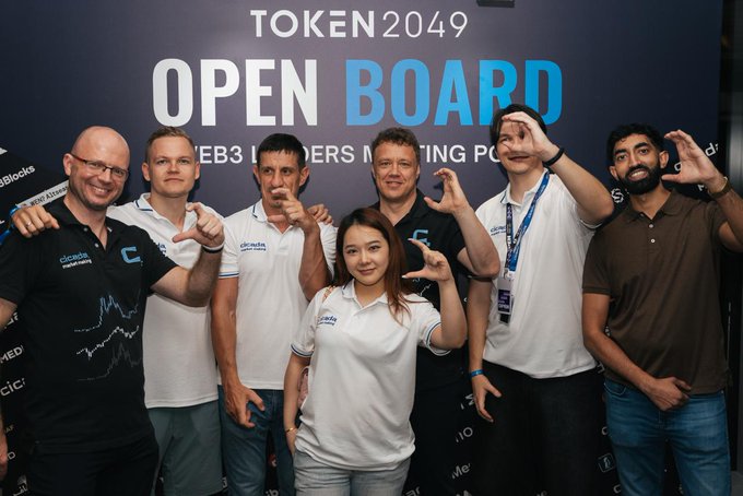 A group photo of seven people posing together at the TOKEN2049 event, standing in front of a backdrop reading 'OPEN BOARD' with 'Cicada Markets' shirts visible.