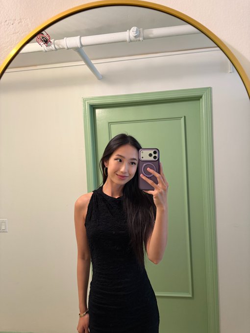 Mirror selfie of a young woman with long dark hair wearing a sleeveless black dress, holding a smartphone in front of a green door and arched gold mirror.