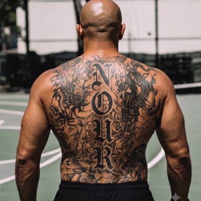 A muscular, bald man's back displays a large tattoo spelling 'NOIR' vertically in ornate lettering.