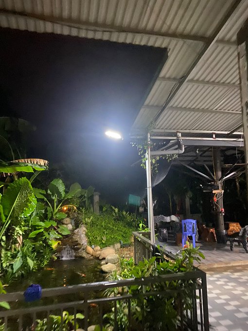 A serene nighttime scene of a covered patio area featuring a small illuminated pond with a fountain, surrounded by lush tropical plants, with a bright light glowing under the corrugated roof and a blue plastic chair visible in the background. The image complements the post's weekend relaxation theme, showcasing a tranquil evening retreat perfect for unwinding from a busy week of market activities.