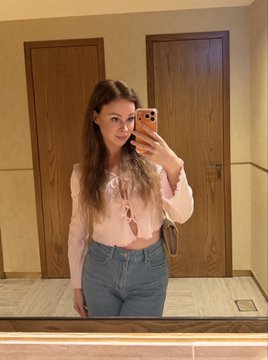 A mirror selfie of a young woman with long brown hair wearing a tied pink long-sleeve top and blue jeans, taken in a room with wooden doors.