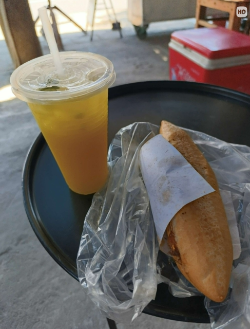 A casual outdoor meal consisting of a plastic cup of iced tea or juice with a straw and a wrapped sandwich or banh mi in a plastic bag, placed on a black tray with a red cooler in the background.