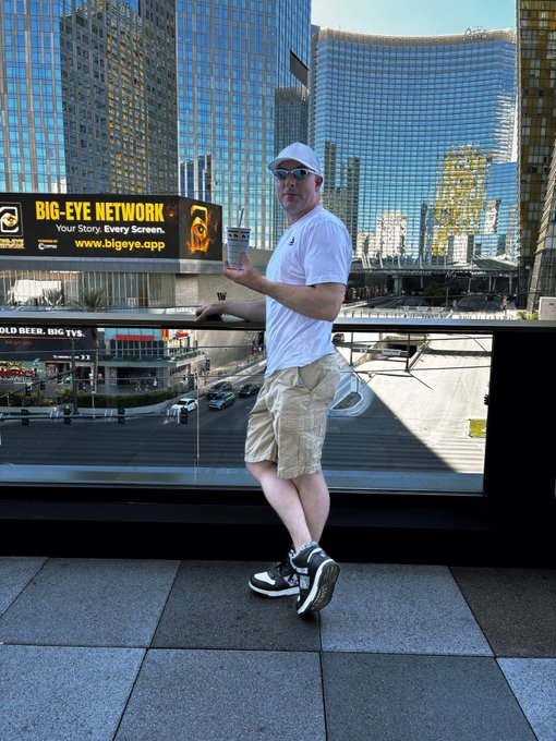 A man wearing a white cap, sunglasses, white t-shirt, and beige shorts stands on a balcony holding a drink, with a 'BIG-EYE NETWORK' billboard visible across the street in a city setting.