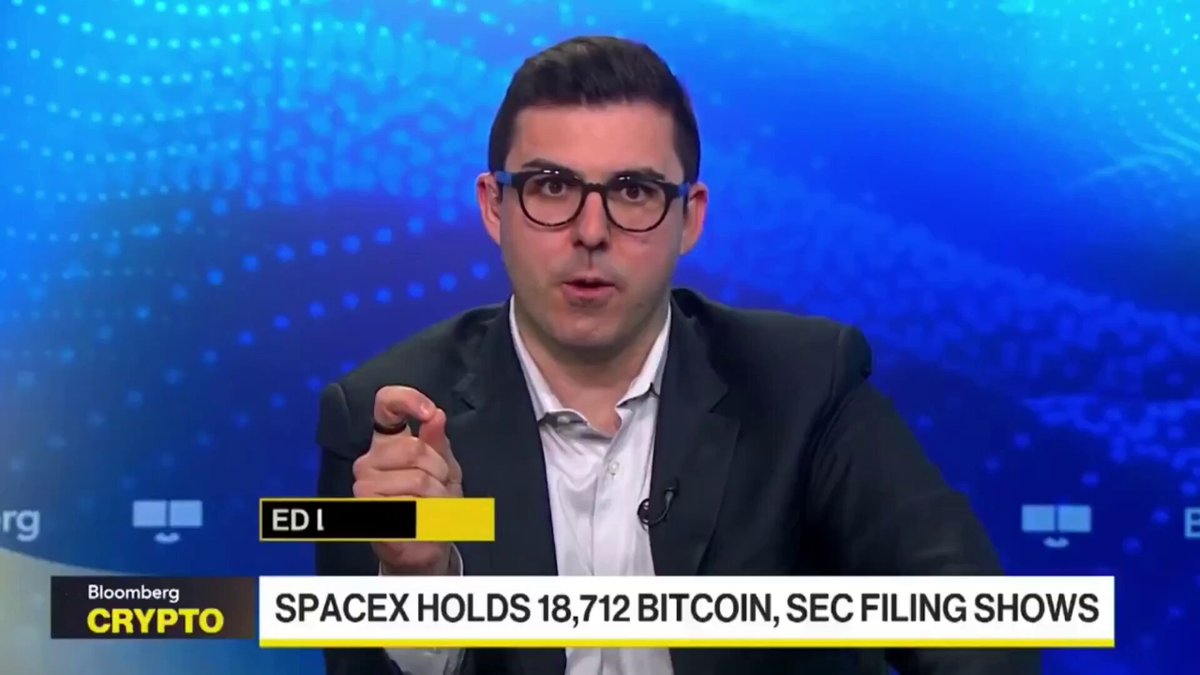 The image shows a man wearing glasses sitting at a desk with his hand extended, possibly gesturing or pointing towards something. The background of the image features a blue wave pattern, which appears to be part of an animated news segment related to Bitcoin. The text in the image reads "HUGE: SpaceX holds 18,712 BTC, sec filing shows," indicating that the company has invested in Bitcoin and is now holding its value as a strategic reserve for excess cash.