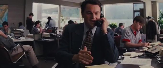 A man in a suit talks on a telephone at a busy 1980s-style trading floor with multiple brokers working at desks in the background.