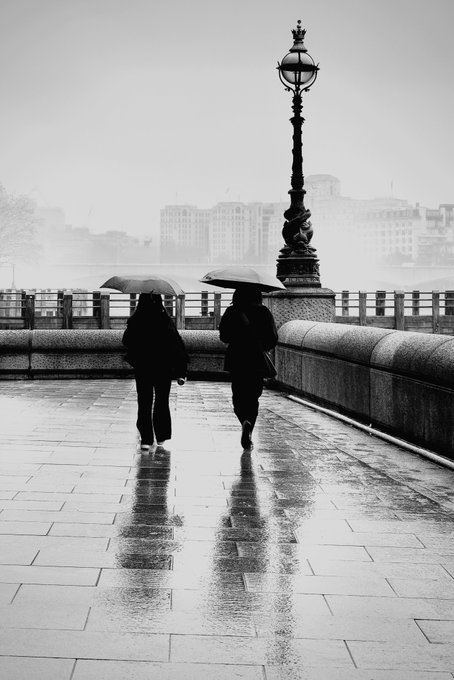 The image features two people walking down a sidewalk with umbrellas, likely due to rain or other wet weather conditions. The scene takes place during the day under an overcast sky, which adds to the mood of the photo. There are no visible texts in the image, and the focus is on the people and their surroundings.