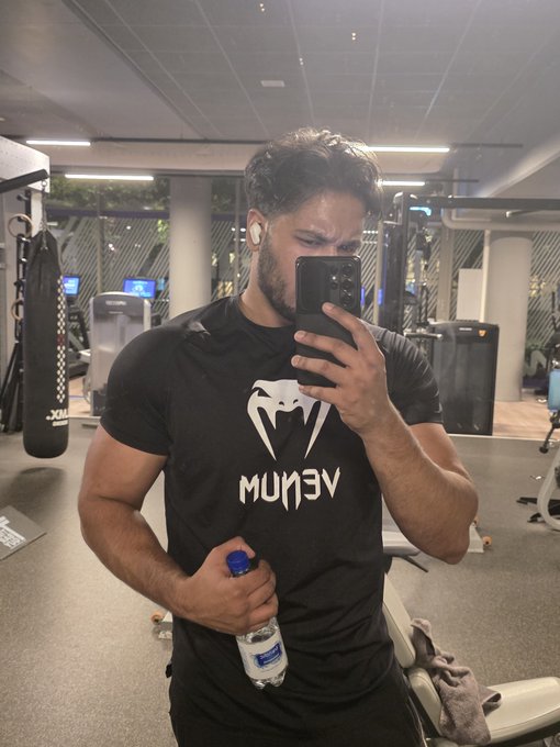 A muscular man takes a mirror selfie at the gym wearing a black **VENUM** t-shirt (with the snake logo and mirrored text) and holding a water bottle and phone. The image captures his post-workout physique, embodying the "Gym builds the body" side of the post's message about combining fitness with crypto investing.
