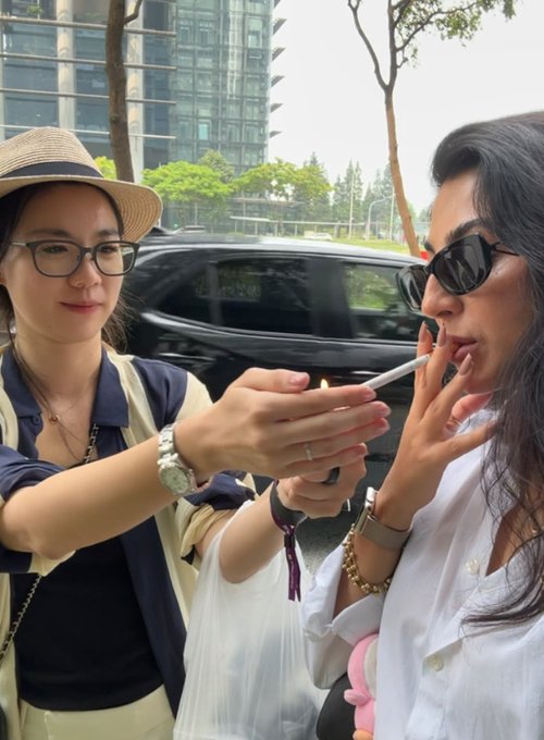 One stylish woman lights a cigarette for another outside near a parked car, with Singapore high-rises visible in the background, suggesting an urban networking moment during the crypto conference.