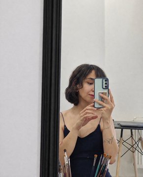 A short-haired woman takes a mirror selfie with her phone, with paintbrushes visible at the bottom of the frame.