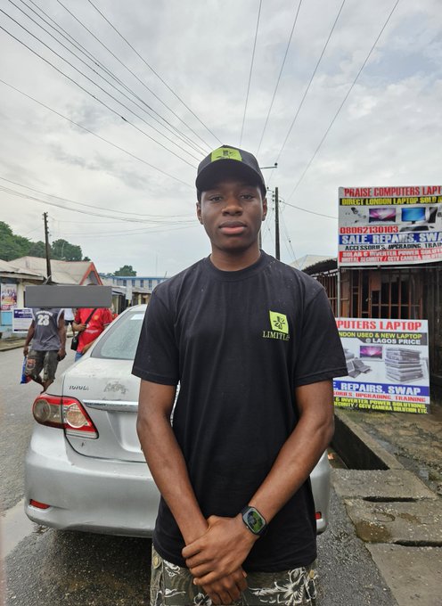 The image features a man standing next to his car with a sign that says "Limitless" above him. The man appears to be wearing a black shirt and camo pants, giving off a hipster vibe. There are other people visible in the background, but they don't seem to be the main focus of the photo.