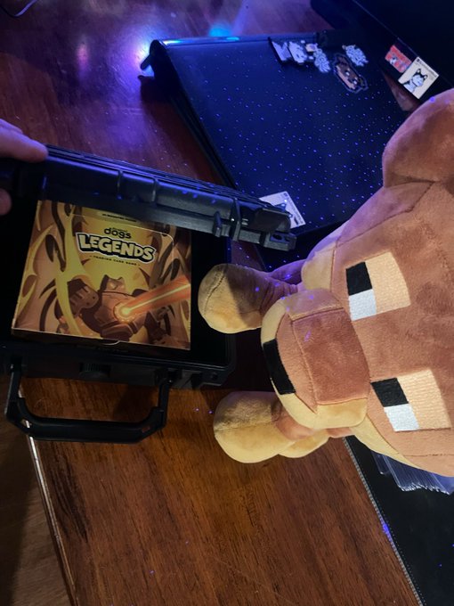 A person's hand opens a case revealing a 'doga LEGENDS' album pack, with a Minecraft character plush toy sitting nearby on a wooden table.