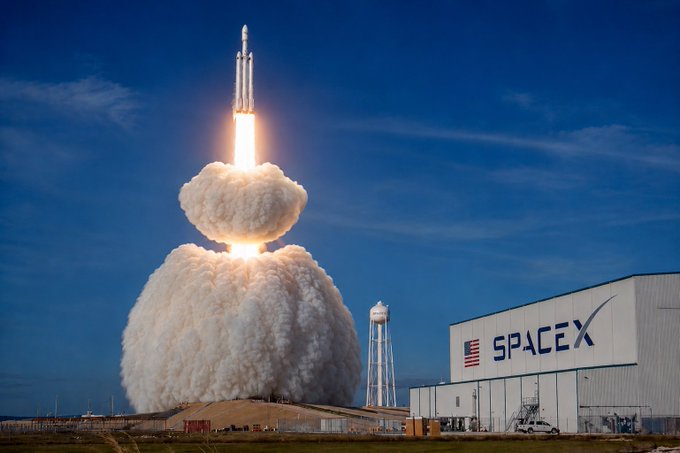 A SpaceX rocket lifts off in a fiery ascent from the launch pad, with a massive plume of smoke billowing beneath it against a blue sky. The "SPACEX" logo and American flag are prominently displayed on the adjacent hangar, tying into the caption "all my bags go to space" by evoking the excitement and ambition of space travel.