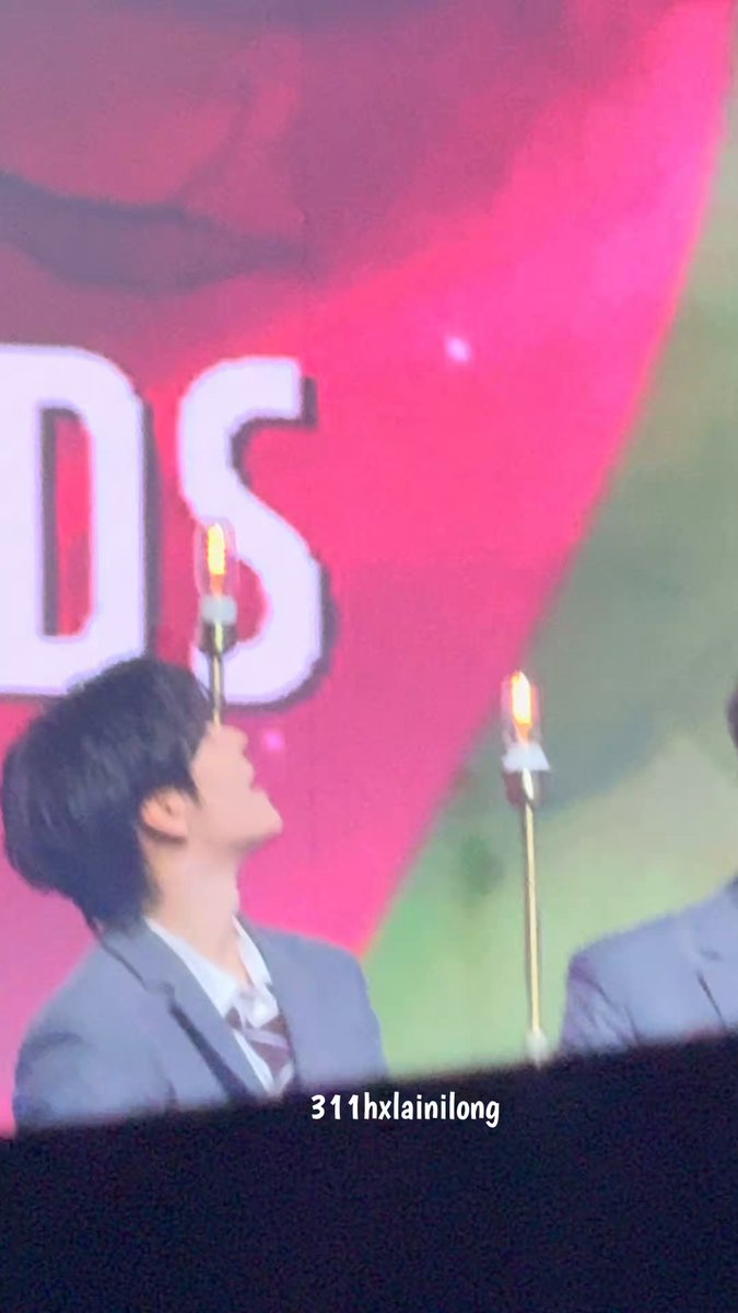 The image features a young man wearing a suit, standing in front of a large screen with a pink background that says "DS" on it. The man appears to be looking up at something or someone above him, possibly an award or recognition. There is also another person partially visible behind the main subject, but their face is not clearly visible due to the angle and distance from which the photo was taken.