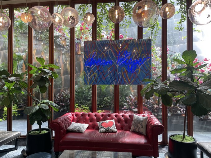 A stylish interior featuring a red tufted leather sofa with decorative pillows, flanked by potted fiddle leaf fig trees, in front of large windows looking into a garden area with a blue neon sign.