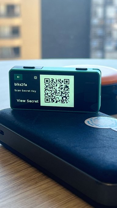 The image shows an electronic device with a QR code displayed on its screen. The device appears to be a laptop computer placed on top of a notebook or a small black case. The QR code seems to contain some sort of secret key, possibly related to Bitcoin or 2FA accounts. The text in the image suggests that this device is used for viewing and managing sensitive information securely.