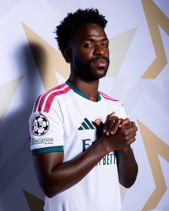 A man modeling a white Arsenal FC away football kit featuring pink shoulder accents, a UEFA Champions League 15 sleeve patch, and the 'Visit Rwanda / Foundation for charity' sleeve sponsor.