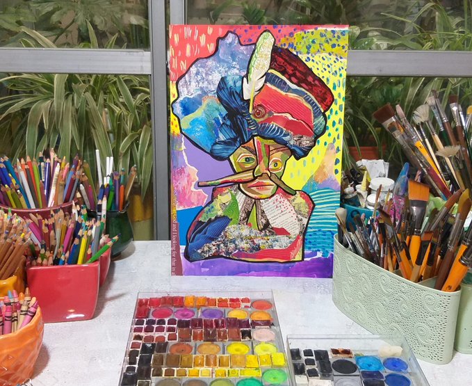 A colorful painting of a face with a long nose and feathered headdress displayed on an easel, surrounded by containers full of colored pencils and a watercolor palette.