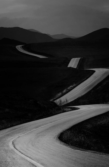 A black-and-white photograph of a winding road snaking through dark, rolling hills under a moody sky.