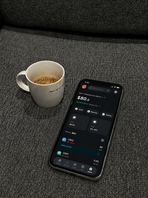 A smartphone displaying a crypto wallet app with a balance of $80.00, alongside a white coffee mug on a dark textured surface.