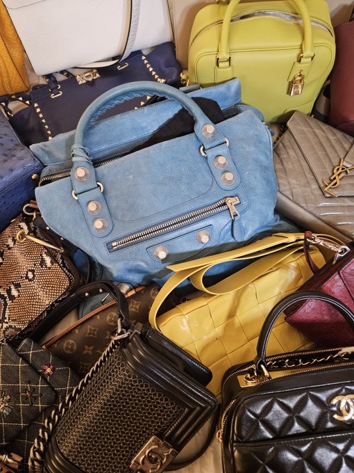 A collection of luxury designer handbags piled together, including a blue Balenciaga-style bag, a yellow Hermès Birkin, and what appears to be Louis Vuitton and Chanel pieces.