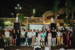 A large group of people stand together outdoors at night in front of a stage with speakers and tropical trees.