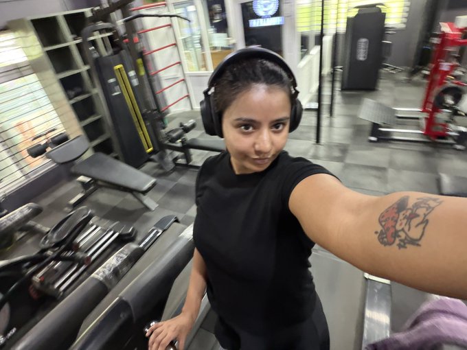 A young woman wearing black headphones takes a selfie at a gym, flexing her arm to reveal a tattoo while surrounded by weight equipment. The image ties to the post's fitness motivation, showing a dedicated gym-goer who likely just finished a workout that includes squats to better handle life's challenges.