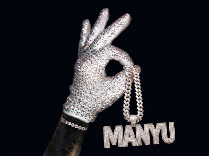 A hand wearing a diamond-encrusted glove holding a large diamond chain pendant spelling 'MANYU', referencing the rapper Manyuu.