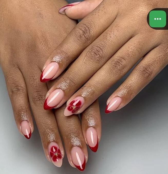A close-up image of hands featuring long, almond-shaped manicured nails painted in a nude pink color with red French tips and red flower designs on two accent nails.