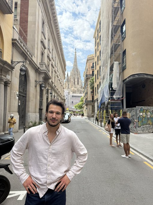A man in a white button-up shirt stands smiling on a city street, with the Gothic spires of Barcelona Cathedral visible in the background between surrounding buildings.