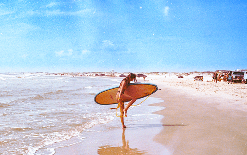 A vintage-style photograph of a woman in a bikini running along a beach shoreline while carrying an orange surfboard, with a clear blue sky and distant beachgoers.