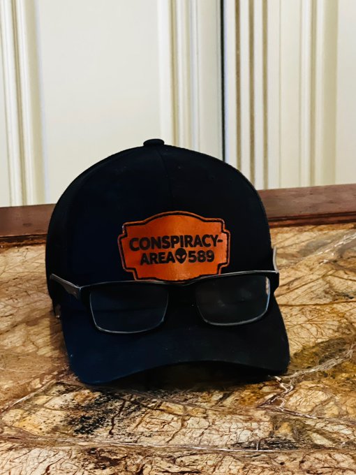 A dark blue baseball cap with an orange leather patch reading 'CONSPIRACY AREA 589' sits on a marble surface with sunglasses resting on its brim.