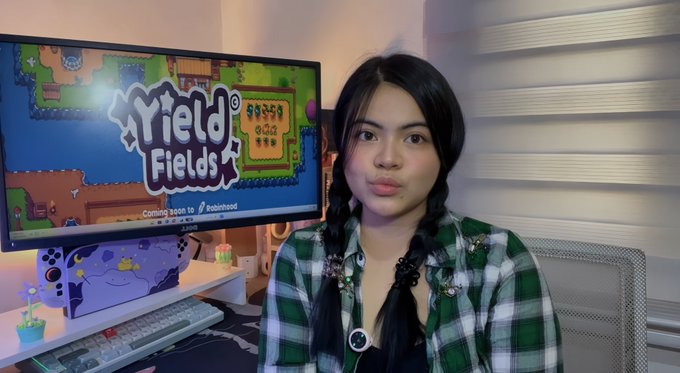 A woman with braided hair in a green plaid shirt sits at a desk with a monitor displaying the game title 'Yield Fields' and 'Coming soon to Robinhood'.