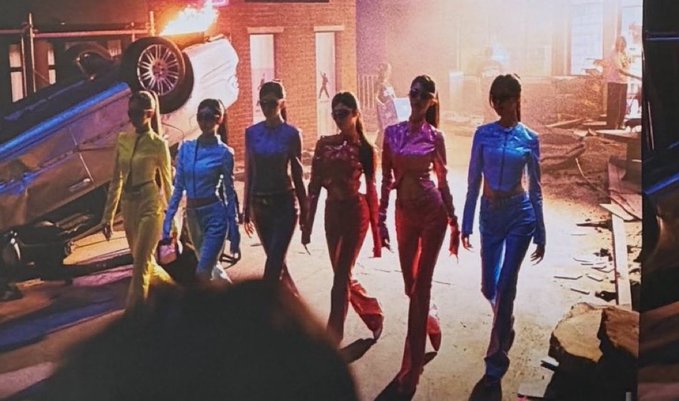 A promotional image showing six women in colorful outfits (yellow, blue, dark blue, red, pink, blue) walking together in a dramatic scene with an explosion and flipped car in the background.