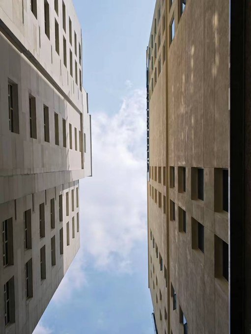 Looking up between two tall stone-colored apartment buildings against a blue sky with wispy clouds, the image conveys a sense of being back outdoors after being busy—"am back" vibes as the poster returns to share with friends.