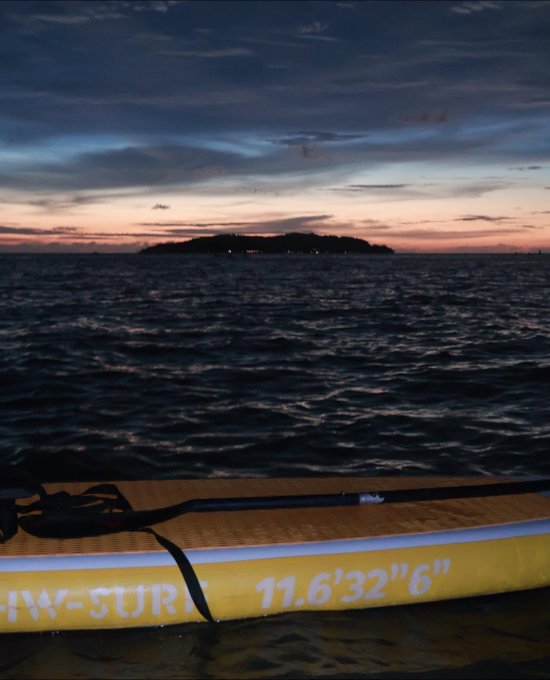 A tranquil evening seascape view from a paddleboard (marked '11'6"32"6"') looking out toward a small island silhouetted against a colorful sunset sky.