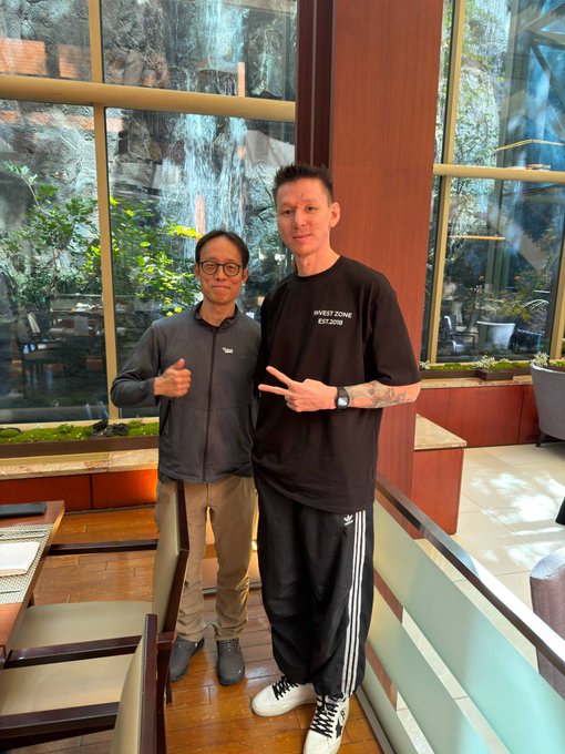 Two men posing for a photo in front of a large window with a waterfall view; the taller man on the right wears a black "SWEET ZONE EST.2008" t-shirt and makes a peace sign, while the shorter man on the left gives a thumbs up.