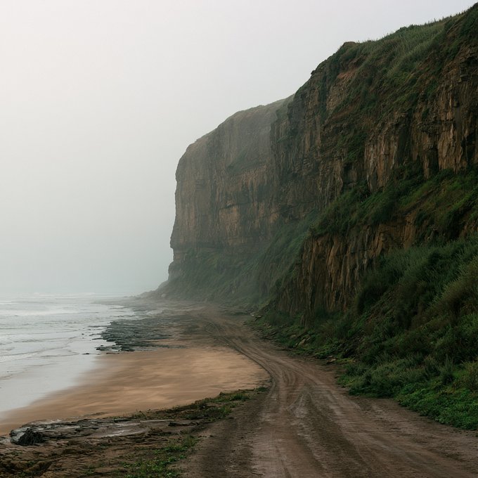 A misty, atmospheric photograph of a rugged coastal cliff face descending to a sandy beach alongside a dirt path.