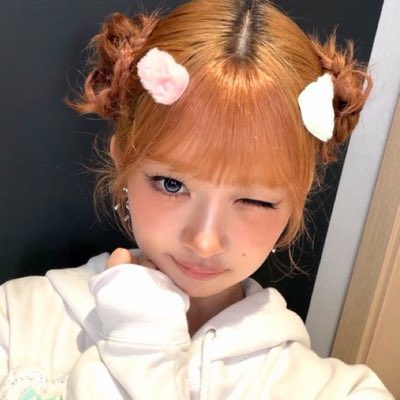 A close-up selfie of a young woman winking at the camera with her hand under her chin, sporting orange hair styled with cat ear buns, pink bows, and fluffy cheek stickers.
