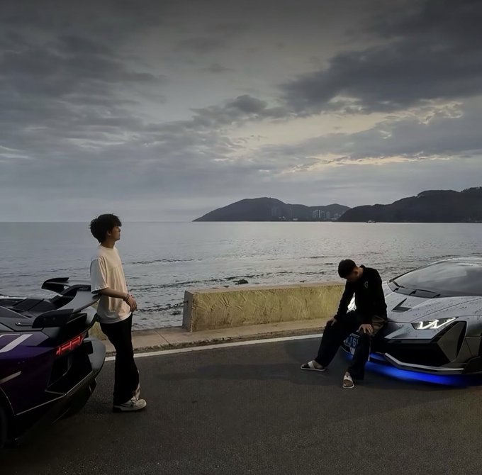 Two young men pose with high-performance sports cars (one purple, one grey) along a coastal road at dusk, with the ocean and a distant headland visible in the background.